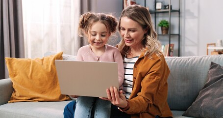 Wall Mural - Portrait of happy nice Caucasian family daughter and mother spending time together at home using laptop online. Mom and cute small child girl tapping on computer surfing internet, leisure concept