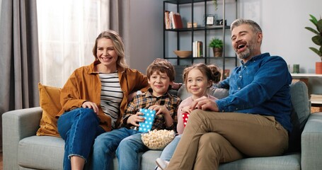 Wall Mural - Caucasian joyful happy Caucasian family resting at home watching TV together sitting on couch with popcorn and laughing. Mom and dad with small son and daughter eating popcorn watching comedy movie