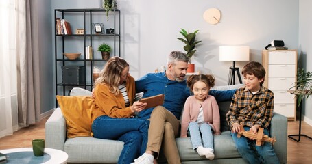 Wall Mural - Caucasian cheerful lovely family parents and small kids together at home. Little son boy playing while parents sitting with small daughter watching at tablet screen, videochatting, family time concept