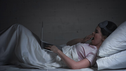 Wall Mural - tired freelancer in eye mask using laptop and yawning in bed at night