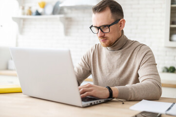 Serious focused determined young bearded businessman or freelancer in glasses and casual clothes sitting at kitchen table, typing on laptop, working on project, researching, writing email to client