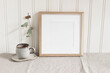 © tabitazn - Empty square wooden picture frame mockup. Cup of coffee on linen table cloth. Dry eucalyptus taped on white wooden wall background. Home office concept. Scandinavian interior design.