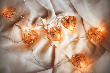 A glowing garland with heart-shaped bulbs glows with a warm light and lies on a white soft fabric, creating comfort and romance. Valentine's day. Wedding.
