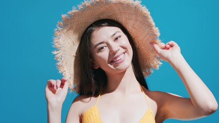 Wall Mural - Young brunette asian woman in yellow crochet swimsuit hiding behind straw hat puts it on her head and smiles for a camera against blue background | Sunscreen commercial concept