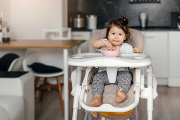 The child himself eats food at home in the kitchen. Cute little Asian girl eats fruit puree. The child is sitting in a high chair. The girl eats with a spoon. The child himself eats food 