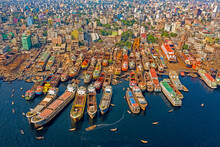 Aerial View Of Several Boats Under Construction Docked Along Buriganga River In Keraniganj With City Skyline In Background, Dhaka, Bangladesh.