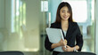 © bongkarn - Businesswoman holding document file and smiling to camera while standing in office room