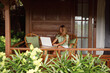 Young woman  freelancer working on laptop computer sitting at table on wooden balcony of country house surrounded with greenery