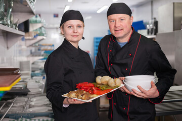 Portrait of two confident cooks standing with cooked dish in restaurant kitchen ..