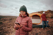 © StratfordProductions - Happy young woman in winter clothing using smartphone during camping in mountain hill while man building tent in background