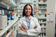 © StratfordProductions - Cheerful young african american female pharmacist wearing labcoat with folded hands standing in chemist near shelves