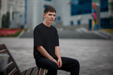 Black t-shirt mockup on a young guy sitting on a bench on a blurred background in the park, stylish clothes for design presentation.