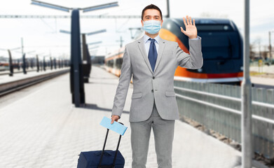 Wall Mural - business trip, health and people concept - businessman in mask with travel bag and ticket waving hand over train on railway station on background