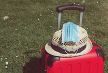 Light Blue Surgical Mask On Luggage And A Hat, In A Beautiful European Green Background. Face Mask Is Compulsory For Travelers, New Normal.