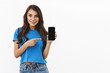 © Cookie Studio - Advertisement, technology. Happy smiling caucasian woman in basic blue t-shirt, holding smartphone, pointing finger at mobile screen, promote game, phone application, white background