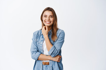 Wall Mural - Portrait of smiling beautiful woman touching her face with natural make up and looking cheerful at camera, standing against white background