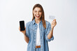 © Cookie Studio - Online shopping. Cheeky young woman winking at you, recommending mobile banking app, showing empty smartphone screen and plastic credit card, white background