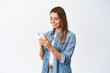 © Cookie Studio - Portrait of young blond woman chatting on social media, holding smartphone and texting with pleased smile, standing against white background