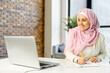 © Vadim Pastuh - Smiling muslim businesswoman taking notes sitting at the desk in modern office space. Young islamic female student wearing hijab and smart casual wear overwrites from the laptop screen into notebook