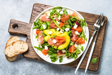 Wall Mural - Green salad with avocado, salad leaves, jamon serrano and tomatoes. Healthy diet lunch. Top view at light stone table.