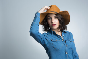 Modella bianca con i capelli neri vestita con una camicia in jeans e un cappello marrone, si tocca la testa con espressione seria  , isolata su sfondo bianco