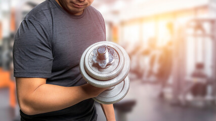 An asian man wear black t-shirt sportswear is lifting a dumbbell with right arm and doing a dumbbell curl for strength training inside fitness club gym. Orange sunlight coming through window.