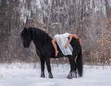 Fototapeta Konie - A beautiful curly-haired girl lies on a Friesian stallion with a long mane