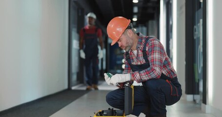 Wall Mural - Bearded middle-aged electrical worker in hardhat protective uniform searching right tools in instruments box. Residential building corridor.