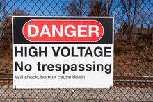 Danger, High Voltage, No Trespassing Sign On A Fence In Front Of A Rail Road. It Lists Possible Health Hazards Including Shock, Trauma And Death Underneath. Train Tracks Seen Blurred In Background.