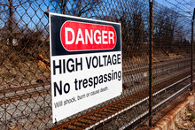 Danger, High Voltage, No Trespassing Sign On A Fence In Front Of A Rail Road. It Lists Possible Health Hazards Including Shock, Trauma And Death Underneath. Train Tracks Seen Blurred In Background.