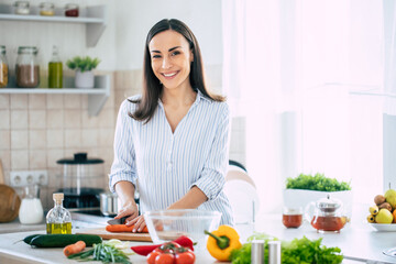 Wall Mural - Cute happy young brunette woman in good mood preparing a fresh vegan salad for a healthy life in the kitchen of her home