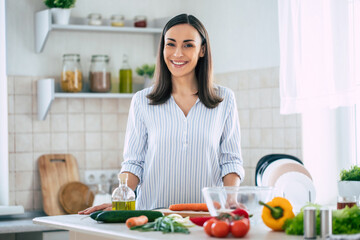 Wall Mural - Cute happy young brunette woman in good mood preparing a fresh vegan salad for a healthy life in the kitchen of her home