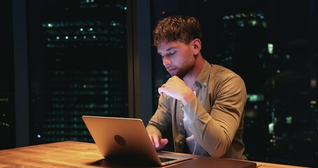 Thoughtful busy young businessman working hard on complicated computer task, developing software application or solving problems online alone till late in modern office with night city view.