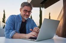 Bearded Man Working Online With Laptop Computer At Home On Terrace. Home Office, Browsing Internet, Tele Working, Video Chat. Portrait Of Mature Age, Middle Age, Mid Adult Man.