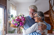 Portrait of couple of two happy and in love seniors or mature and old people holding flowers at home looking outside. Pensioners adult enjoying and celebrating holiday together.