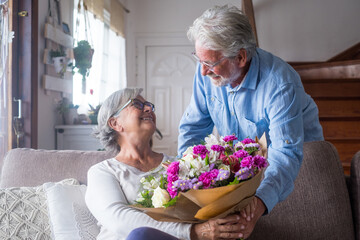 Old man giving flowers at his wife sitting on the sofa at home for the San Valentines’ day. Pensioners enjoying surprise together. In love people having fun.