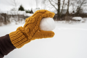 A hand in a yellow mitten holds a snowball, in the yard against the background of a snow-covered vegetable garden. Winter active games and fun. 