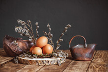 Easter Spring Composition: Eggs In A Basket-nest Of Branches Decorated With Willow Buds. Ceramic Vase, Milk Jug On A Wooden Table. Concept Of A Village Morning And Farming. Dark Background Copy Space.