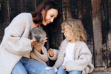 Mother and her daughter play with a little cat by the wooden wall.