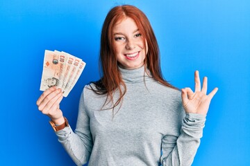 Wall Mural - Young red head girl holding 10 united kingdom pounds banknotes doing ok sign with fingers, smiling friendly gesturing excellent symbol