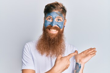 Poster - Young irish redhead man wearing facial mask amazed and smiling to the camera while presenting with hand and pointing with finger.