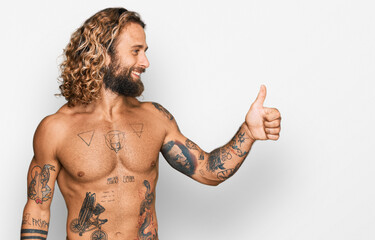 Handsome man with beard and long hair standing shirtless showing tattoos looking proud, smiling doing thumbs up gesture to the side