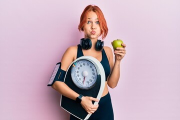 Sticker - Young beautiful redhead woman holding weight machine to balance weight loss puffing cheeks with funny face. mouth inflated with air, catching air.