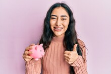 Hispanic Teenager Girl With Dental Braces Holding Piggy Bank Smiling Happy And Positive, Thumb Up Doing Excellent And Approval Sign