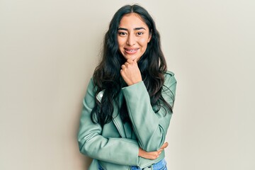 Wall Mural - Hispanic teenager girl with dental braces wearing green leather jacket smiling looking confident at the camera with crossed arms and hand on chin. thinking positive.