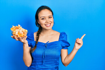 Young hispanic girl holding bowl of potato chip smiling happy pointing with hand and finger to the side