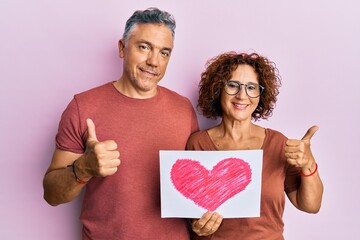 Poster - Beautiful middle age couple holding heart draw smiling happy and positive, thumb up doing excellent and approval sign