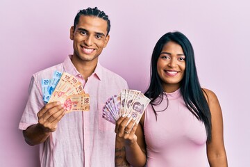 Wall Mural - Young latin couple holding mexican pesos looking positive and happy standing and smiling with a confident smile showing teeth