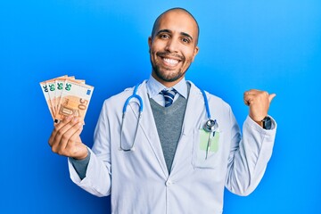 Canvas Print - Hispanic adult doctor man wearing medical uniform holding 50 euros banknotes pointing thumb up to the side smiling happy with open mouth