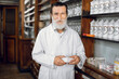 © sofiko14 - Close up portrait of smiling bearded senior Caucasian man pharmacist, posing in vintage drugstore interior, holding box with medicines and looking at camera.